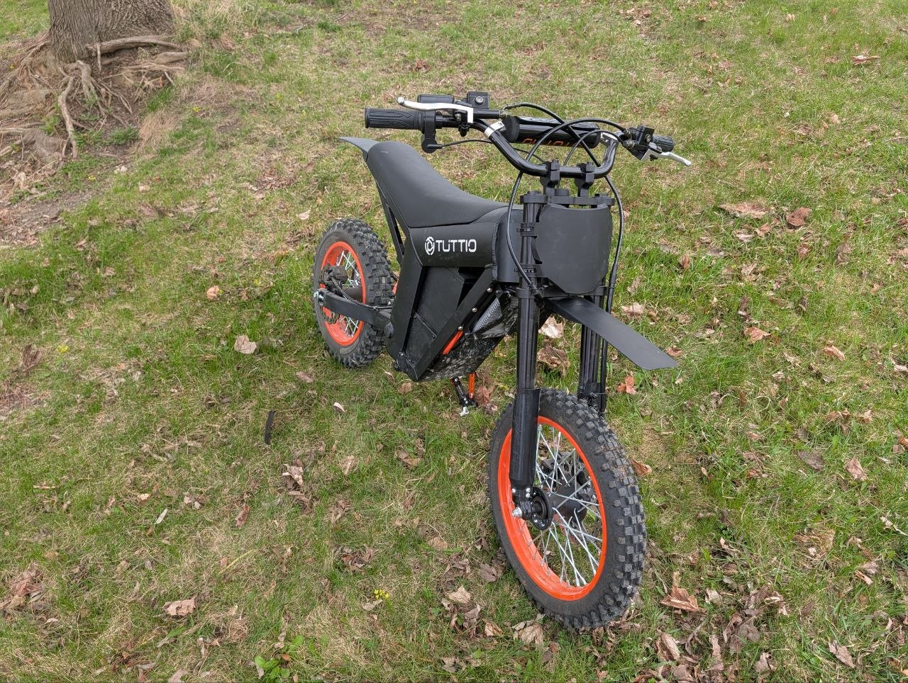Front three-quarter view of the black Tuttio electric dirt bike with bright orange rims, parked on grass next to a tree